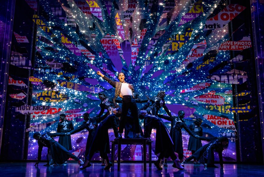 A dancer on stage in a white t-shirt and black pants, in the role of Michael Jackson, with musicians and dancers alongside, in the stage production of MJ the Musical.