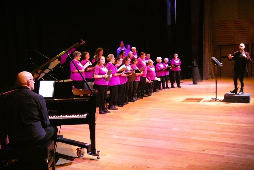 A choir on a wooden stage all dressed in fuscia with a pianist and a conductor. 