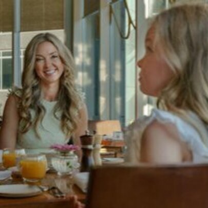 A smiling woman with long blonde hair seated across from a young girl, also with blonde hair. 