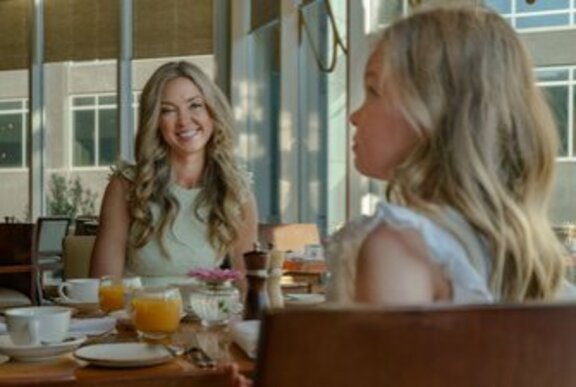 A smiling woman with long blonde hair seated across from a young girl, also with blonde hair. 