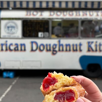 A hand holding a torn open hot jam doughnut in front of a blue and white doughnut van.