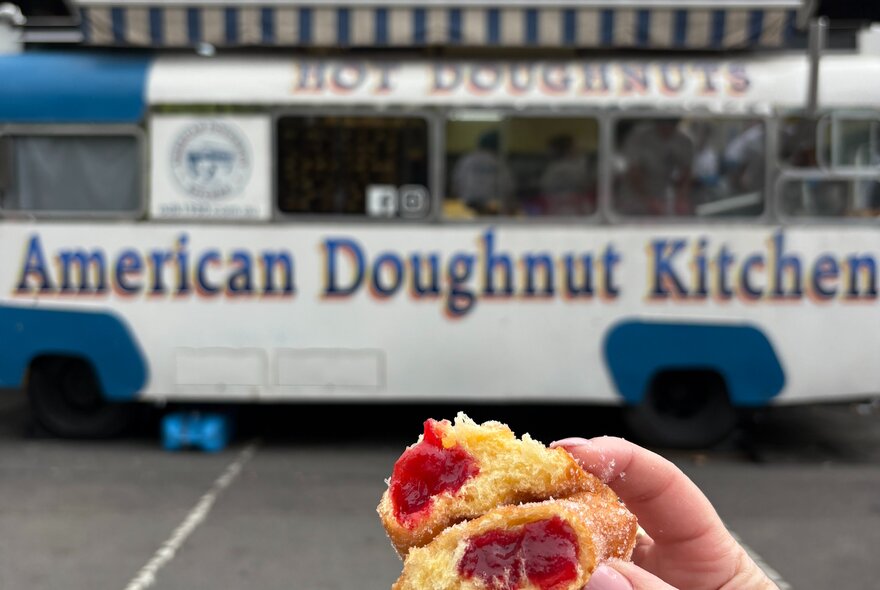 A hand holding a torn open hot jam doughnut in front of a blue and white doughnut van.