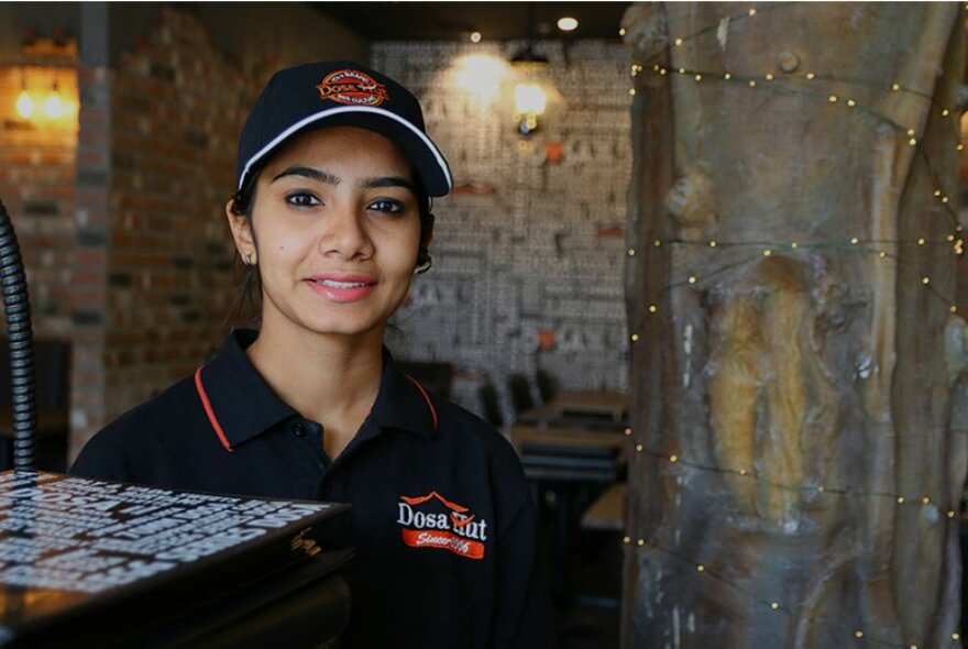 Server in black polo shirt and cap, next to bar, restaurant space at rear.