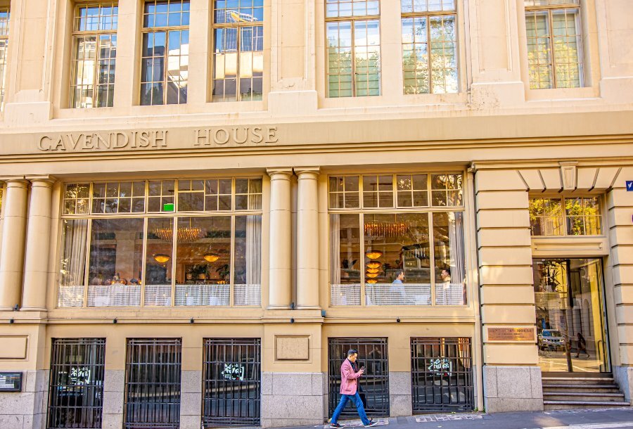 A pedestrian walking past a tall sandstone building with Cavendish House written on the side