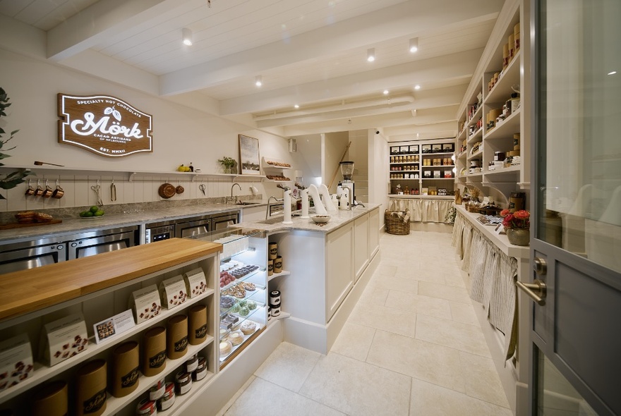 The brightly lit interior of a Mork Chocolates shop showing a white interior, display shelves and a long service counter on the left side. 