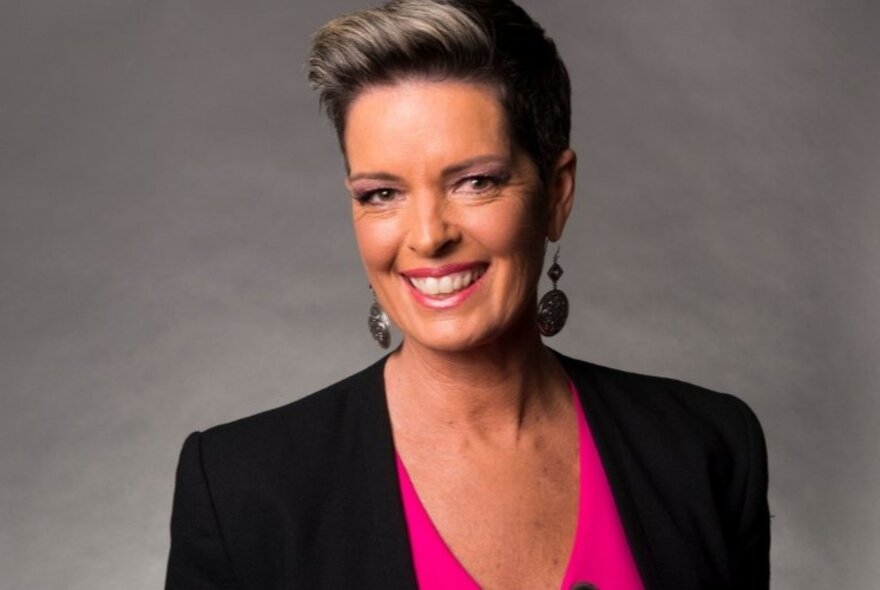 Head and shoulder portrait photograph of journalist Tracey Holmes smiling, wearing a black jacket and pink top underneath, with dangling circular earrings and short hair brushed back off her face.