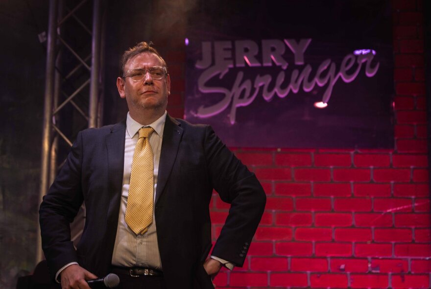 A man in a suit and yellow tie on stage, with a sign that says Jerry Springer on the wall behind him.