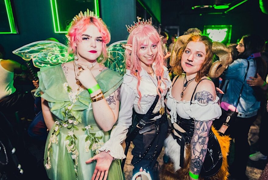 Three women dressed as elves and fairies, with neon green lights in the background.