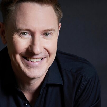 Close-up portrait of the face of conductor Benjamin Northey, a middle-aged man with short dark hair, smiling in a dark open neck shirt.