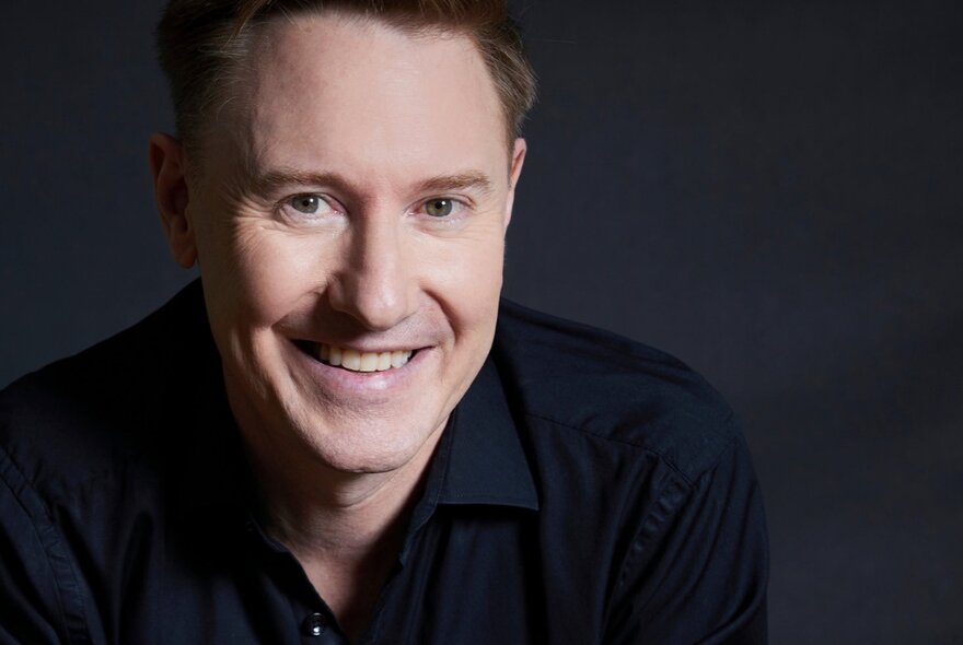 Close-up portrait of the face of conductor Benjamin Northey, a middle-aged man with short dark hair, smiling in a dark open neck shirt.