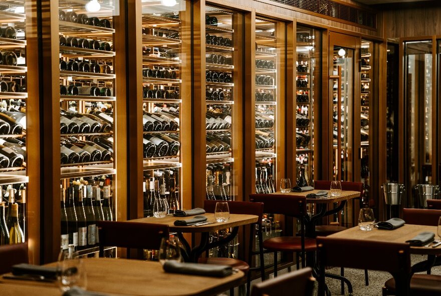 A full wall of timber-lined cabinets filled with wine bottles, behind empty restaurant tables and chairs.