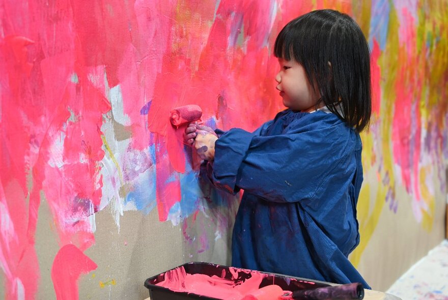 A child in an art smock painting with a small roller on a paper wall. 