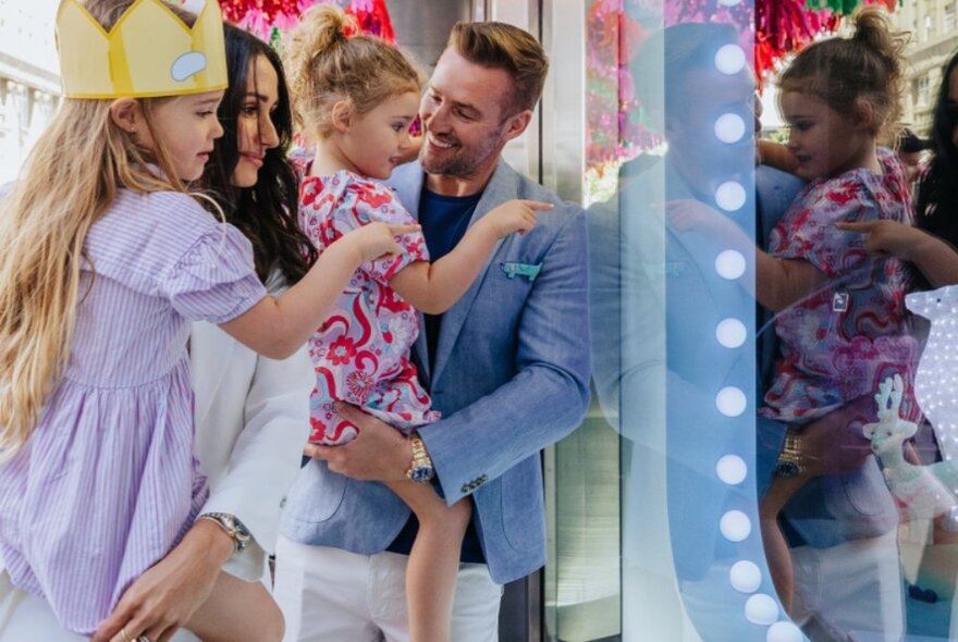 A smiling family of four, looking into the Myer Christmas windows, both young children pointing while being held in their parent's arms.