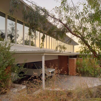 The Pettigrew House, a historic two-storey mid-century residence designed by Australian architect Robin Boyd, with a carport on lower level, an upper level with a large wall of windows, surrounded by leafy green trees.