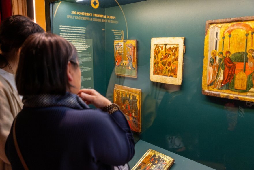 Visitors examine religious wooden icon panels on display on a green wall at the Hellenic Museum.