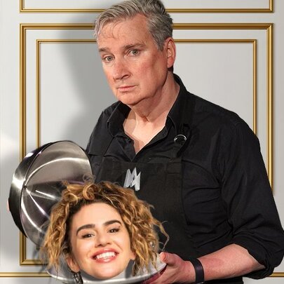 A man in a black button-down shirt and apron stands against a white wall with gold geometric trim, lifting a silver serving cloche to reveal a smiling woman's head resting on a matching platter.