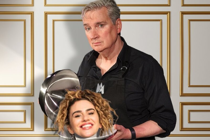 A man in a black button-down shirt and apron stands against a white wall with gold geometric trim, lifting a silver serving cloche to reveal a smiling woman's head resting on a matching platter.