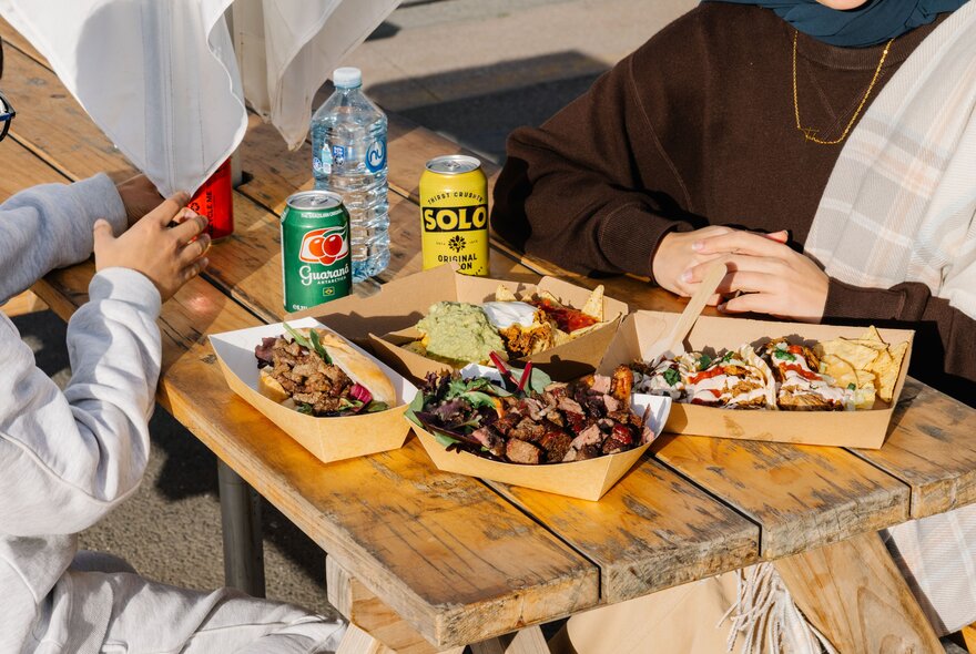 Two people seated at an outdoor rustic wooden picnic table featuring casual street food or food truck meals served in eco-friendly paper trays with cans of soft drink.