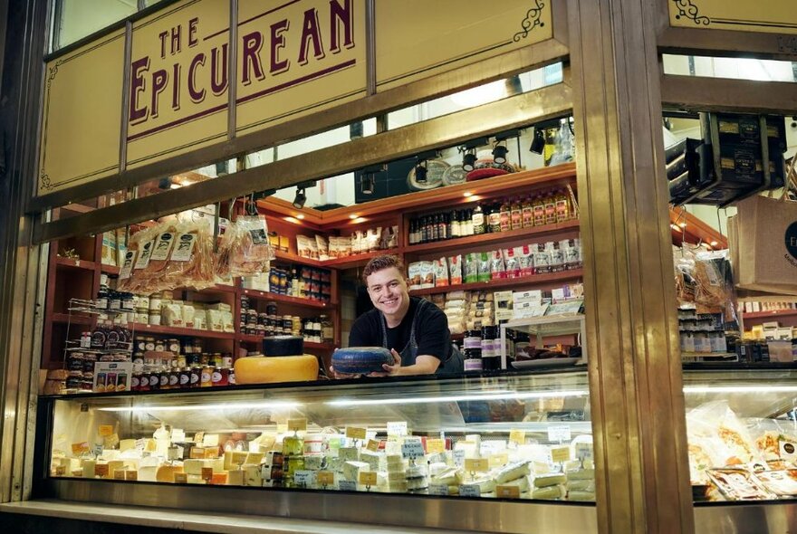 A person serving at a delicatessen stall at Queen Victoria Market, leaning over a large glass display cabinet filled with many different cheeses, with a sign above the counter with the words THE EPICUREAN.