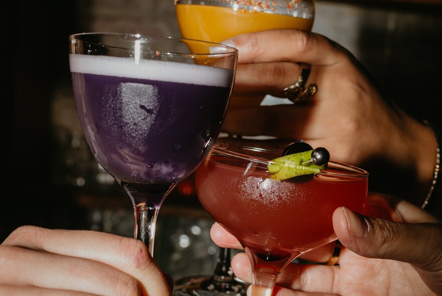 Hands of three people clinking glasses of three different coloured cocktails. 