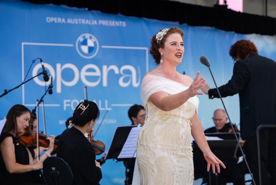 An opera singer dressed in white performing with an orchestra in front of an Opera for all sign. 