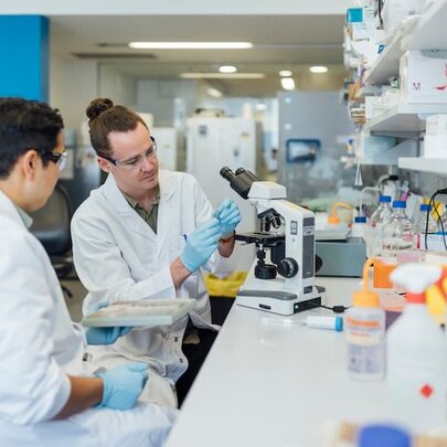 Two white-coated scientists seated in a laboratory with equipment and ingredients.