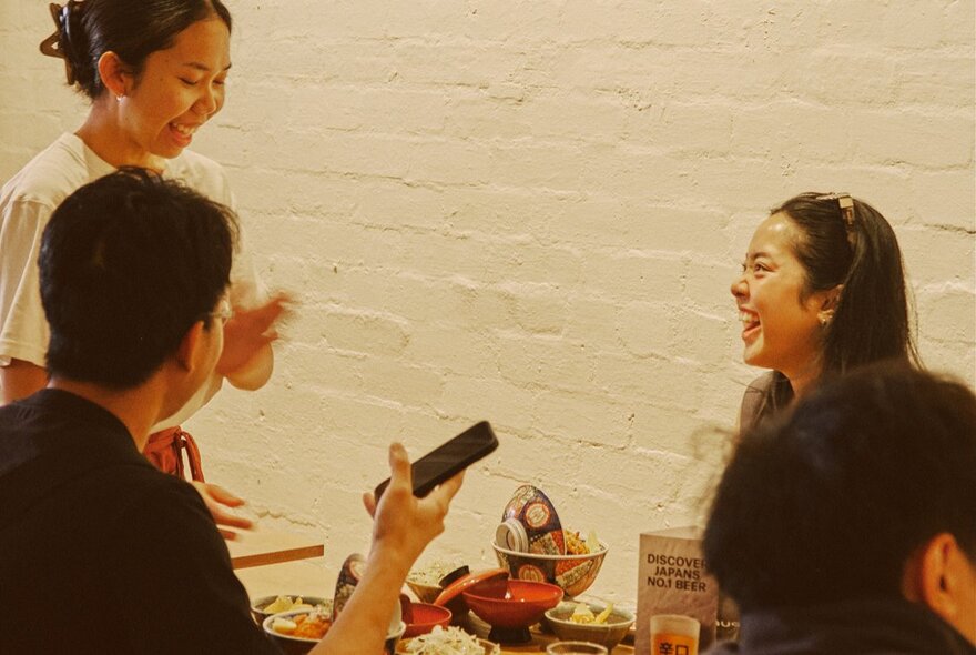 Waitress serving two diners in a Japanese cafe with painted white brick walls.