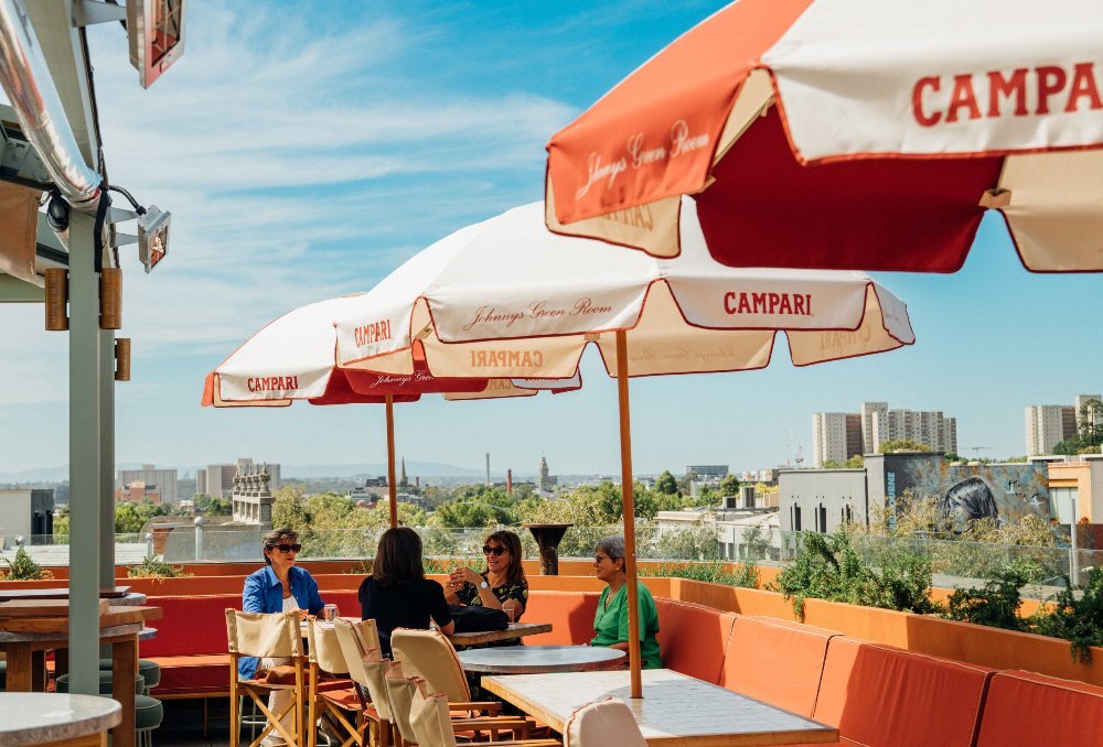 A group of lovely ladies are sitting on a rooftop bar under sun umbrellas with a city skyline view in the background.