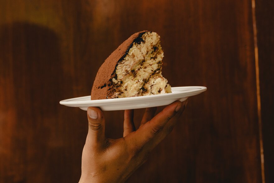 A slice of traditional dome-shaped Florentine ice cream and cake dessert, served on a small white plate being held up by a hand.