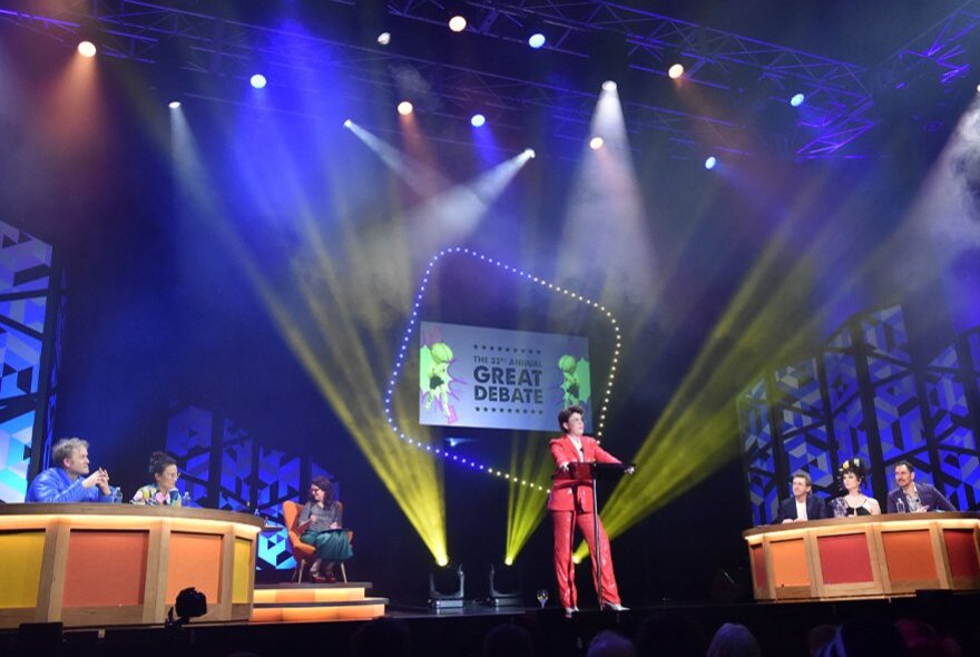 A comedian standing at a lectern and performing on a stage, with two debating teams on either side of the stage seated behind desks.