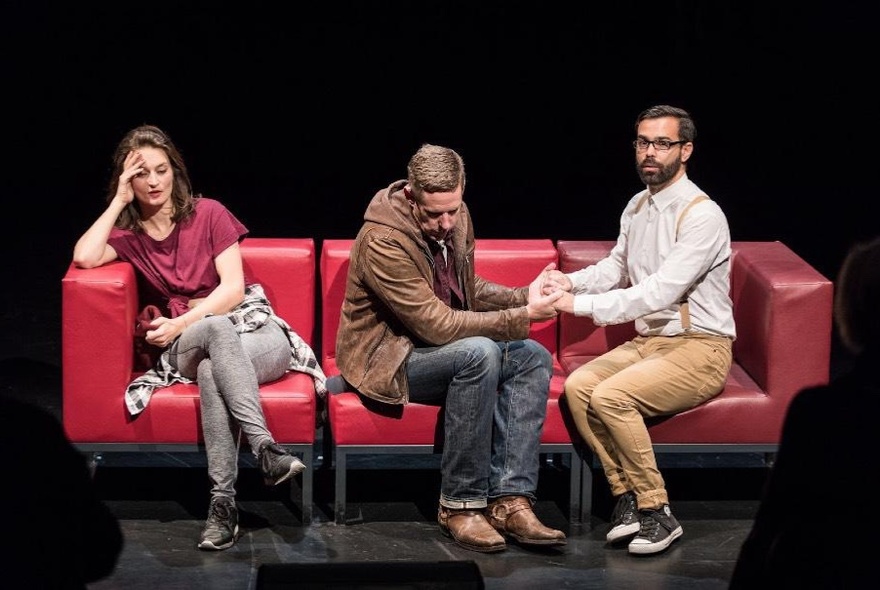 Three improv actors seated on a red leather couch on a stage, the woman on the far left sitting by her self, the two men next to her are facing each other and holding hands.