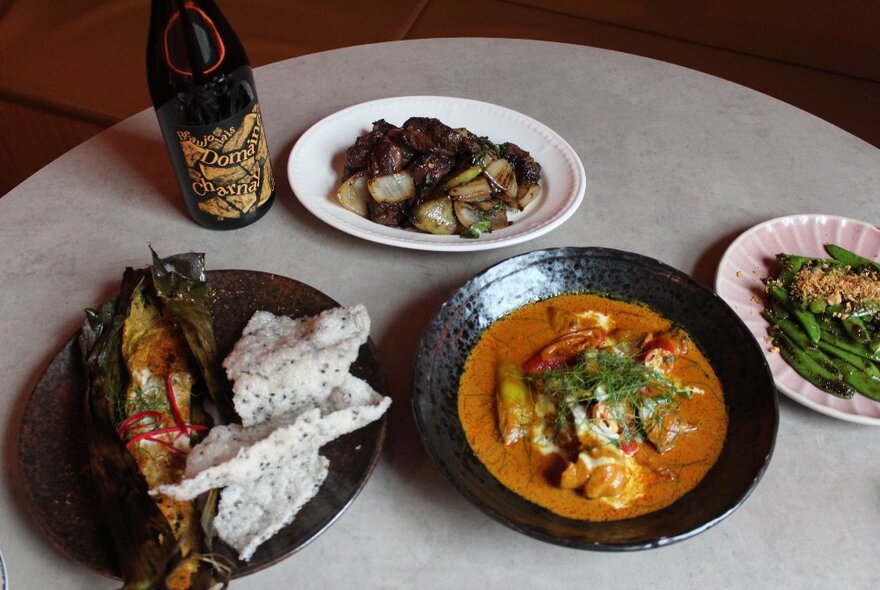 Four dishes of thai-style cuisine on a marble table with a bottle of wine alongside.