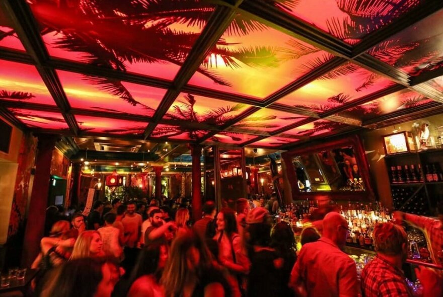 A crowd of people at a bar socialising with the ceiling above them painted in sunset colours and with silhouettes of palm fronds.