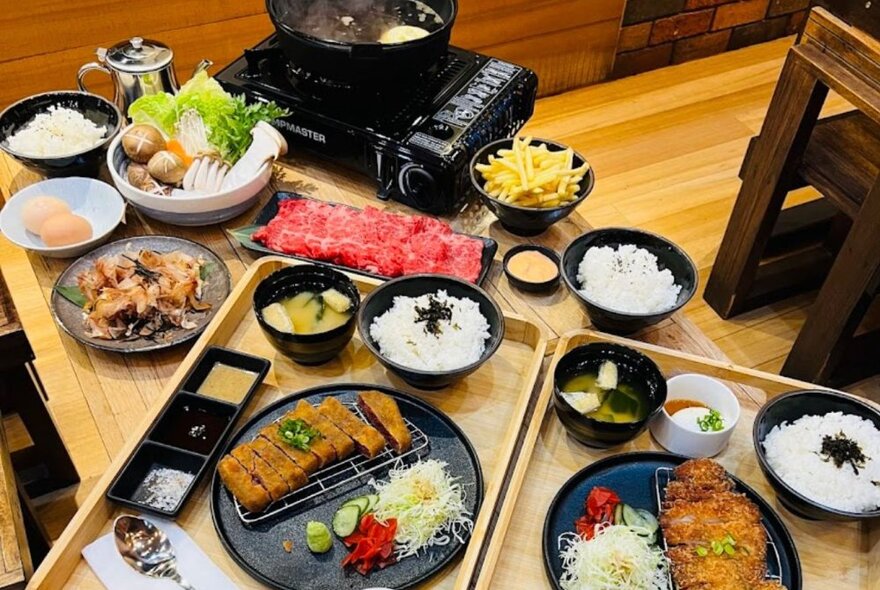 A table filled with different sized plates and bowls of Japanese food, with a small hot plate in the corner. 