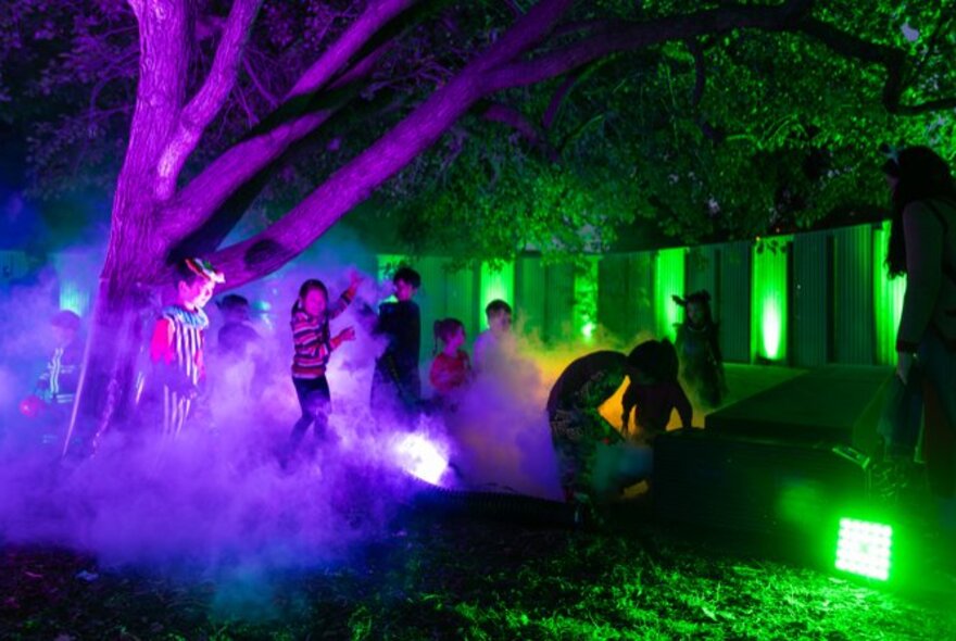 Kids exploring a spooky area with illuminated trees and ground fog with purple and green lights. 