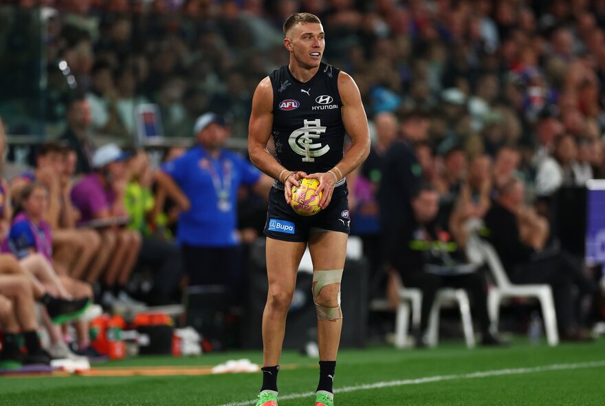 A Carlton AFL football player holding the ball near the boundary during a match, the blurred stadium crowd in the background.