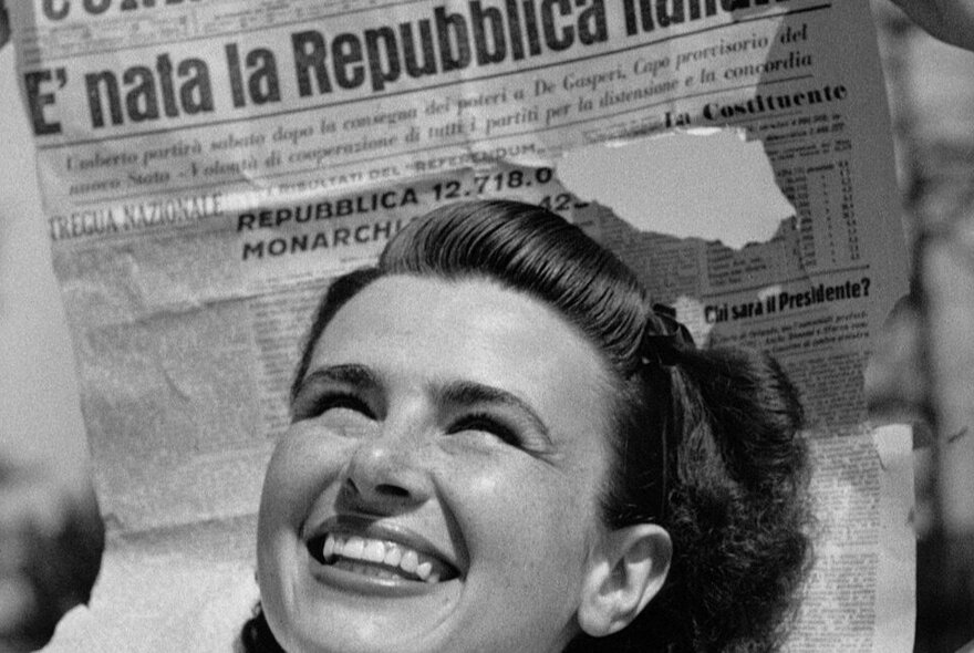 A black and white image of a woman from the 40s with a beaming smile in front of an Italian newspaper. 