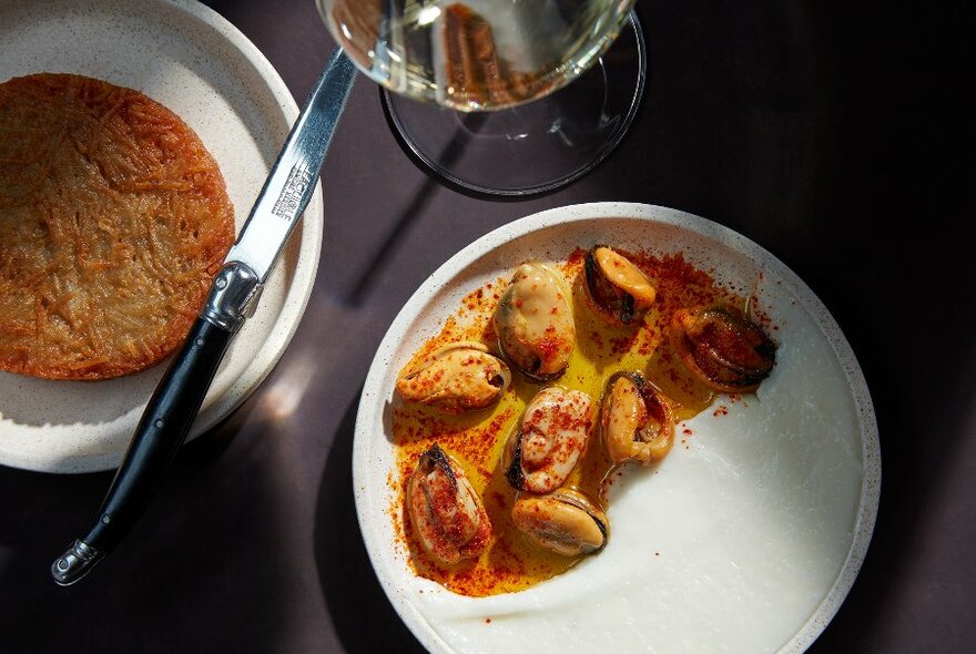 Two plates of food on a dark table with a glass of wine. 