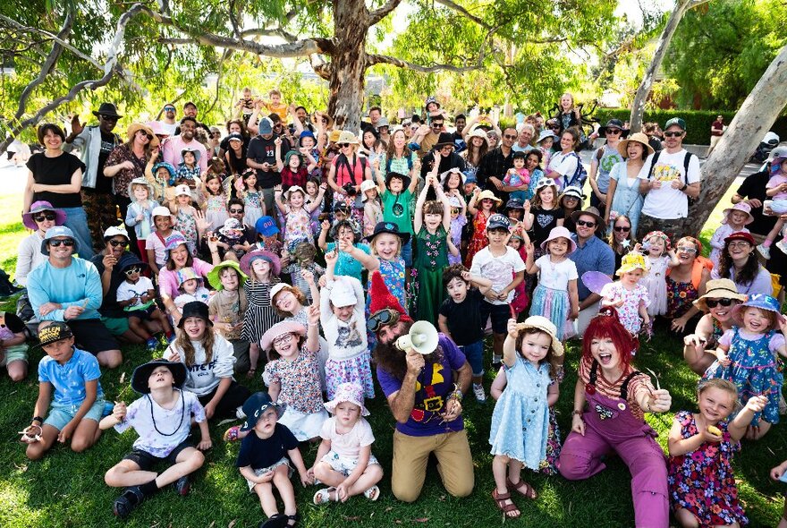 A large crowd of families with young children seated on the grass under a tree, wearing summer hats.