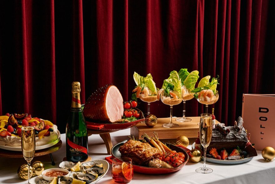 A Christmas buffet presented on a white-clothed table in front of a red velvet curtain, including a ham, prawn cocktails and wine.