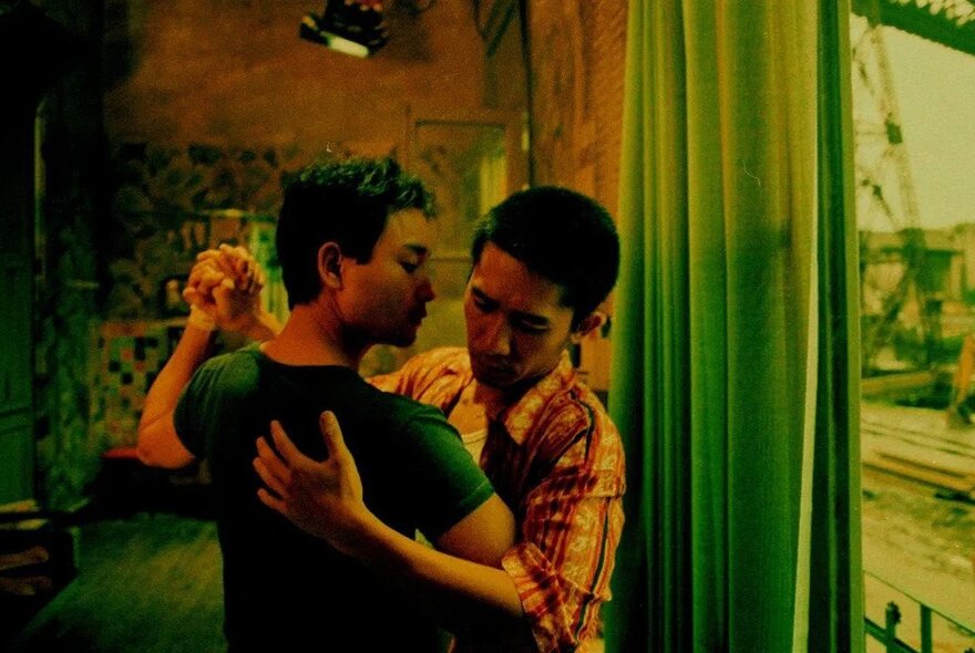 A still from an Asian movie showing a male couple dancing arm-in-arm in front of a window.