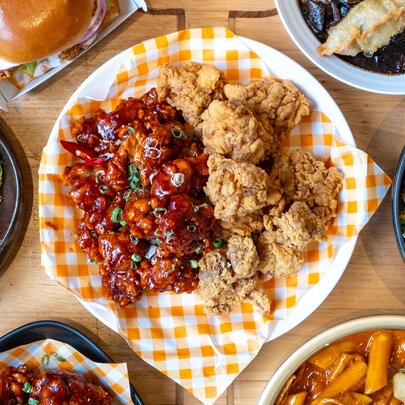 A plate of Korean fried chicken on an orange and white checked napkin, other plates of food around it.