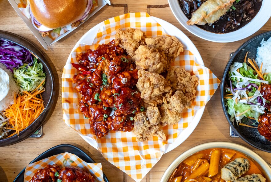 A plate of Korean fried chicken on an orange and white checked napkin, other plates of food around it.