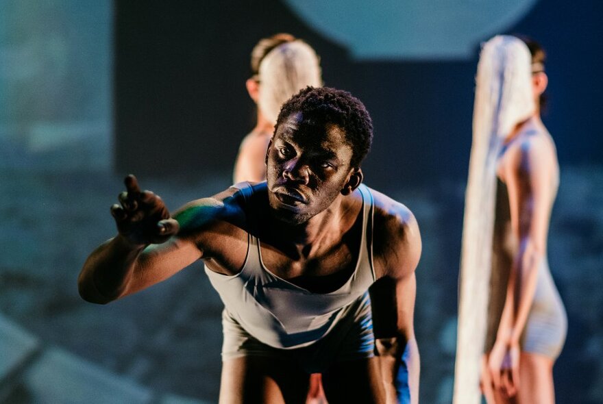 A dancer on stage bent forward, peering at something, with two others in the background with their faces covered by a long piece of material.
