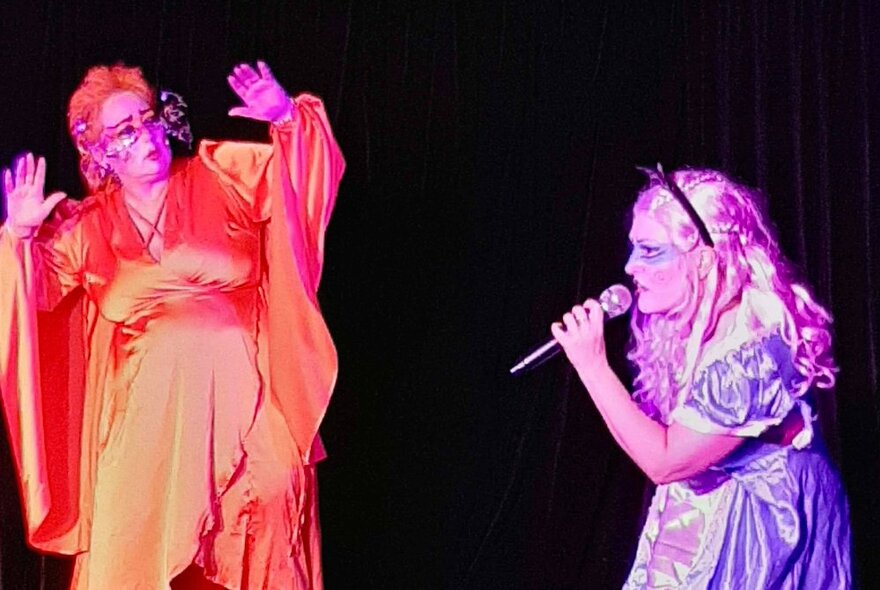 Performers on stage with coloured lights cast on them as one sings into a microphone and they other holds up their hands. 