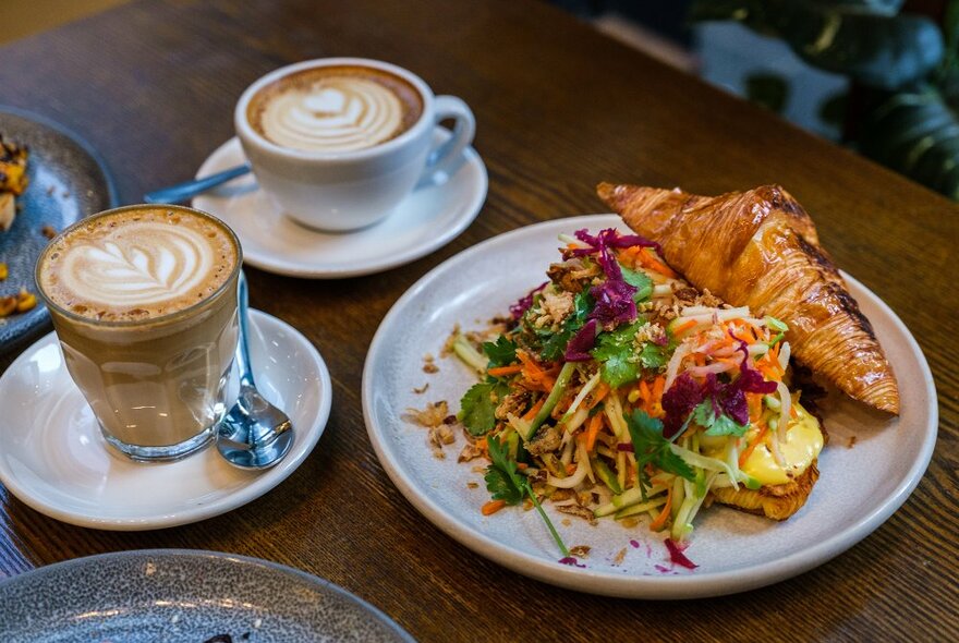 A croissant filled with slaw and two coffees on a cafe table.