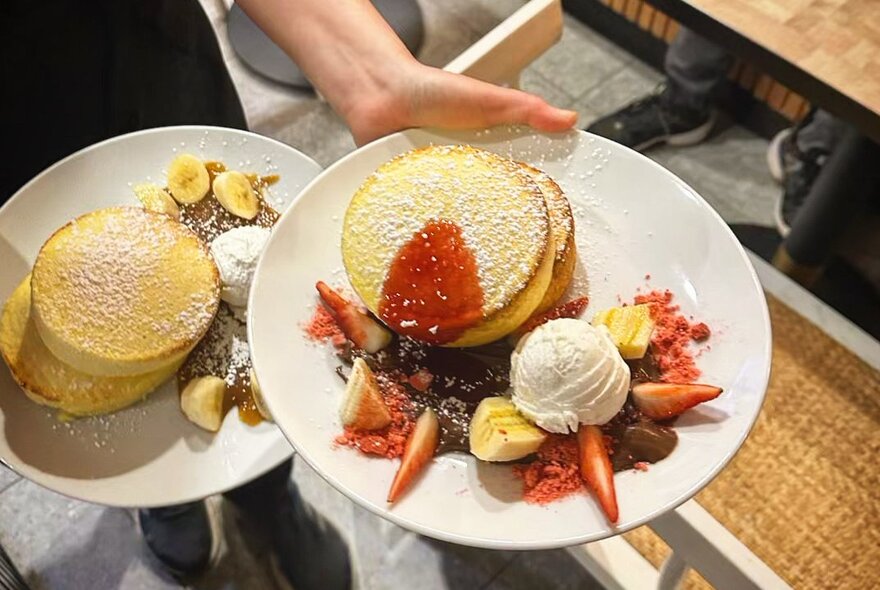 Hands holding two plates of fluffy souffle pancakes with berries and ice-cream.