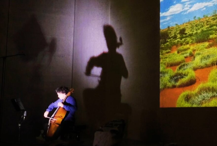 A live musical performance featuring a cellist creating a dramatic shadow effect against a wall while a scenic landscape is projected on a large screen behind them.
