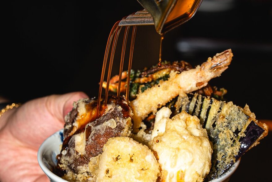 A hand holding a shallow bowl of tempura with a dark sauce being poured over the top.