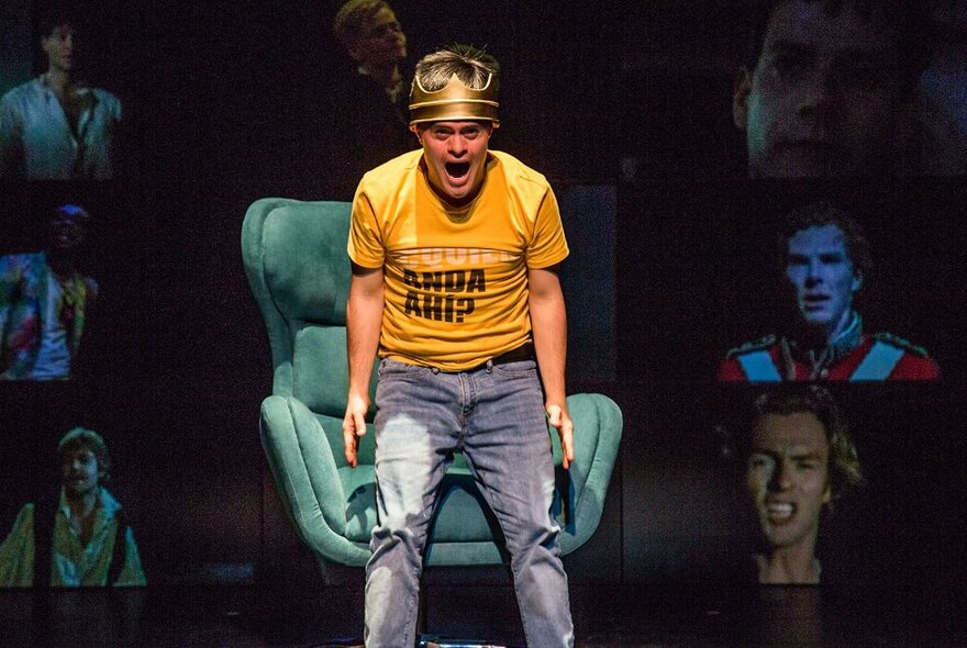A performer on stage in a yellow t-shirt and gold crown, standing up from a teal armchair, screaming, with images of people projected on a screen behind him.